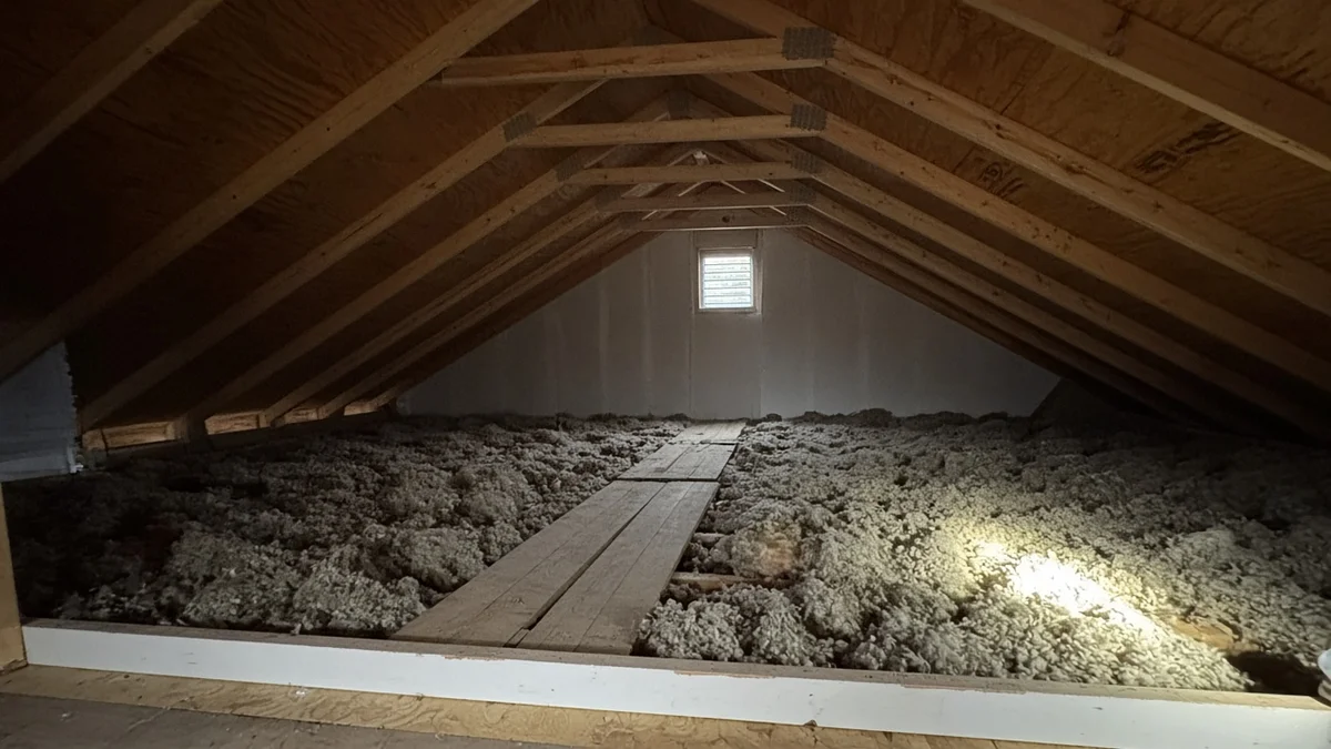 Flashlight shining on blown-in insulation between the roof trusses of a residential attic, with a plank walkway down the middle.